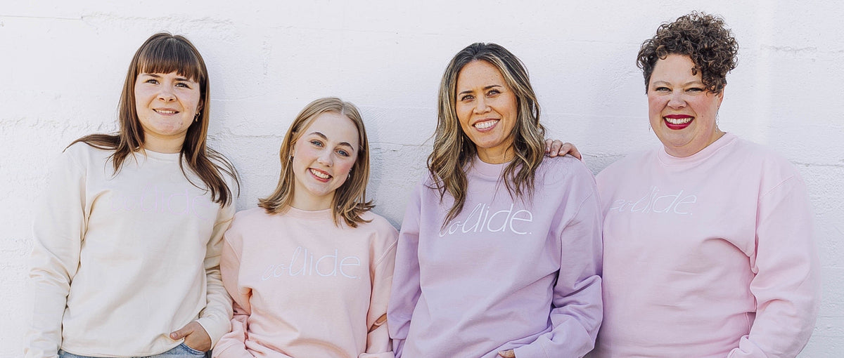 Four people standing next to each other, wearing crew neck sweatshirts in peach, vanilla, lavender, and pink colors, all with a white logo, paired with jeans and casual shoes.